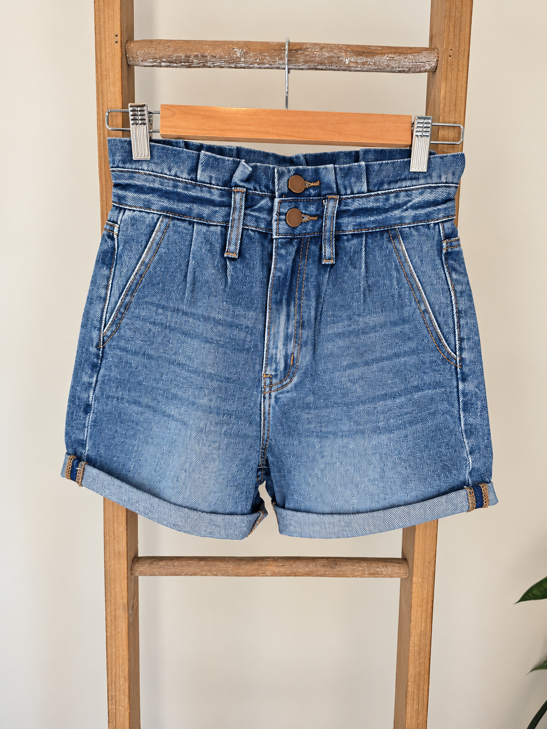 Light blue denim shorts with a high waist, pleated front, and rolled cuffs, hanging on a wooden hanger from a wooden ladder against a light beige wall.