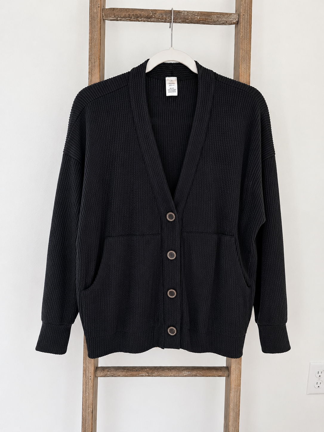 A black ribbed knit cardigan sweater with a V-neck, button closure, and front pockets, hanging on a white hanger against a white wall with a wooden ladder behind it.