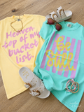Two graphic t-shirts laid flat on a wood floor, styled with white slide sandals, sunglasses, a gold necklace, and bracelets. The left shirt is pale yellow with pink text reading 'Heaven.