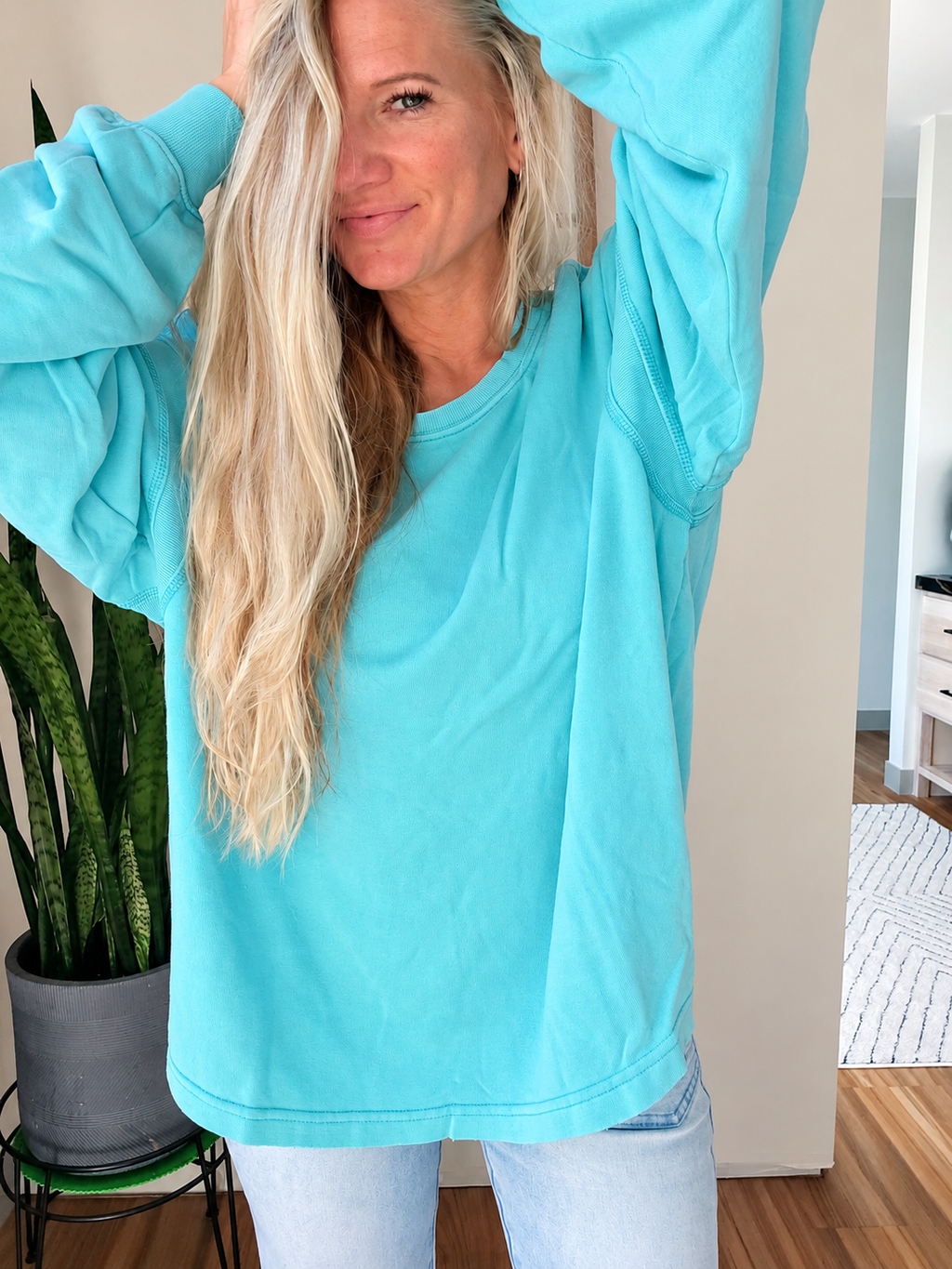 A woman wearing an oversized light blue sweatshirt and light wash jeans, standing indoors with her arms raised in a home setting.