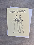 A square greeting card with a textured cream-colored front and a white back, featuring a hand-drawn black ink sketch of a bride and groom and the text 'AWWW YOU GUNS!!' at the top, displayed on a gray textured surface.