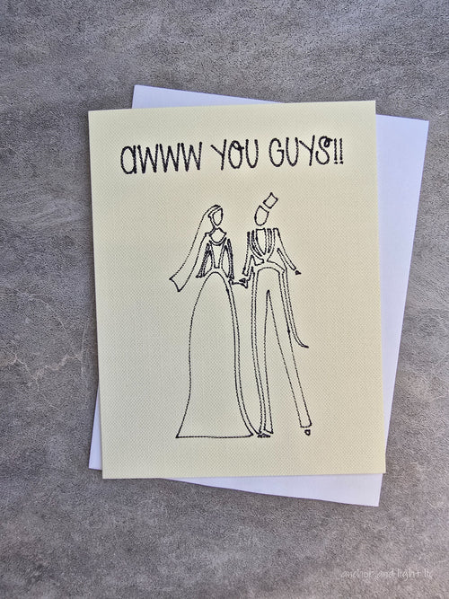 A square greeting card with a textured cream-colored front and a white back, featuring a hand-drawn black ink sketch of a bride and groom and the text 'AWWW YOU GUNS!!' at the top, displayed on a gray textured surface.
