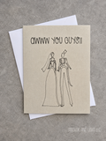 A square greeting card with a kraft-paper style background and black line art of a bride and groom, with the text 'AWW YOU GUYS!!' at the top, shown with a white envelope on a gray textured surface.