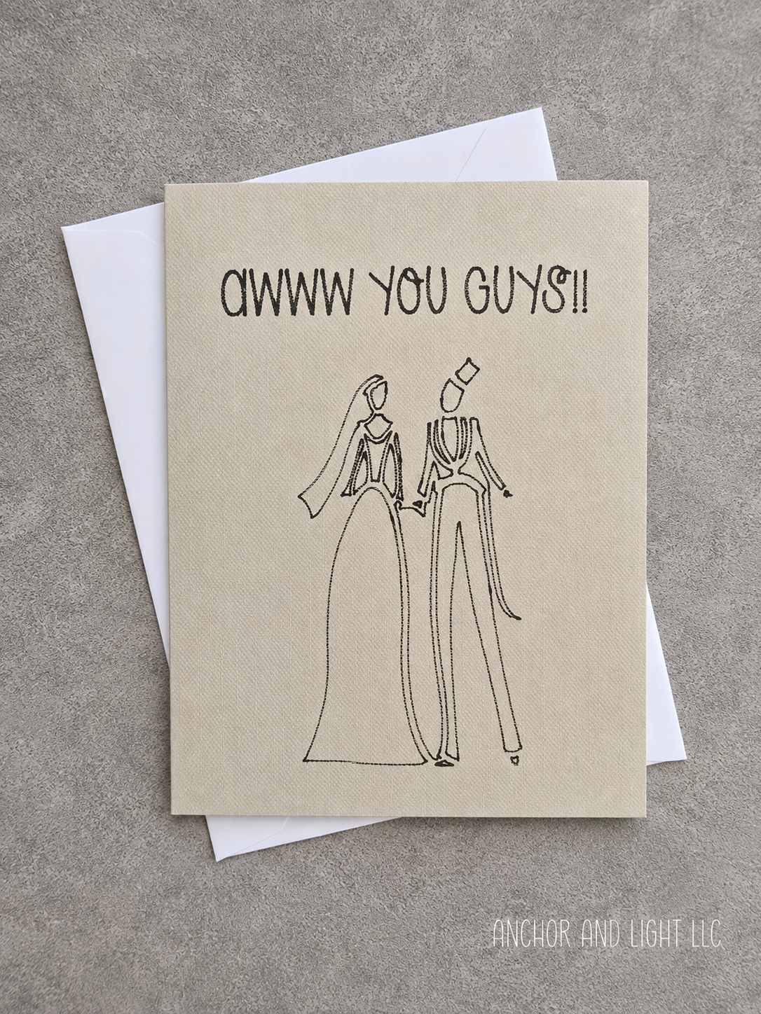 A square greeting card with a kraft-paper style background and black line art of a bride and groom, with the text 'AWW YOU GUYS!!' at the top, shown with a white envelope on a gray textured surface.