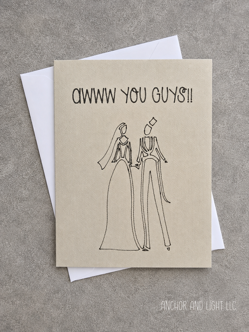 A square greeting card with a kraft-paper style background and black line art of a bride and groom, with the text 'AWW YOU GUYS!!' at the top, shown with a white envelope on a gray textured surface.