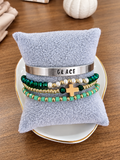 A stack of beaded and metal bracelets displayed on a light blue plush pillow, set on a white plate with a gold rim over a wooden table. The bracelets include a silver cuff engraved with 'GRACE', green and turquoise beads with gold spacer beads, white pearl-like beads, a green and gold cross charm, and dark green beads.