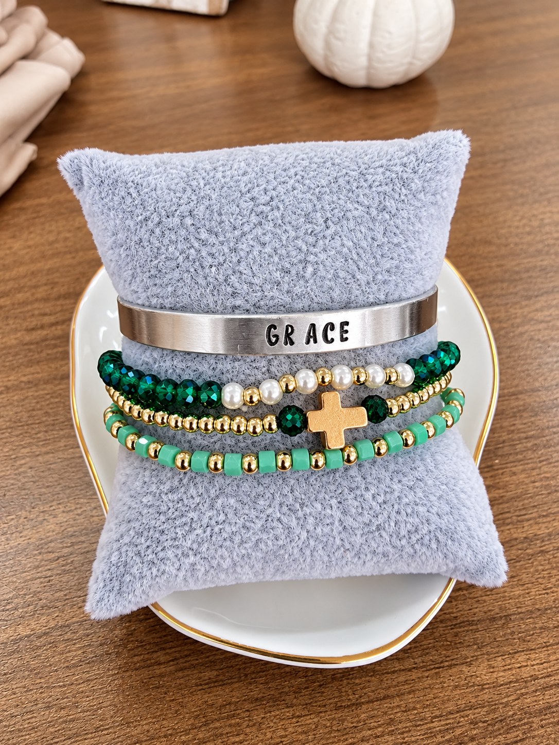 A stack of beaded and metal bracelets displayed on a light blue plush pillow, set on a white plate with a gold rim over a wooden table. The bracelets include a silver cuff engraved with 'GRACE', green and turquoise beads with gold spacer beads, white pearl-like beads, a green and gold cross charm, and dark green beads.