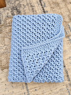A light blue crocheted blanket with an openwork textured pattern, folded neatly on a beige and gray patterned rug.