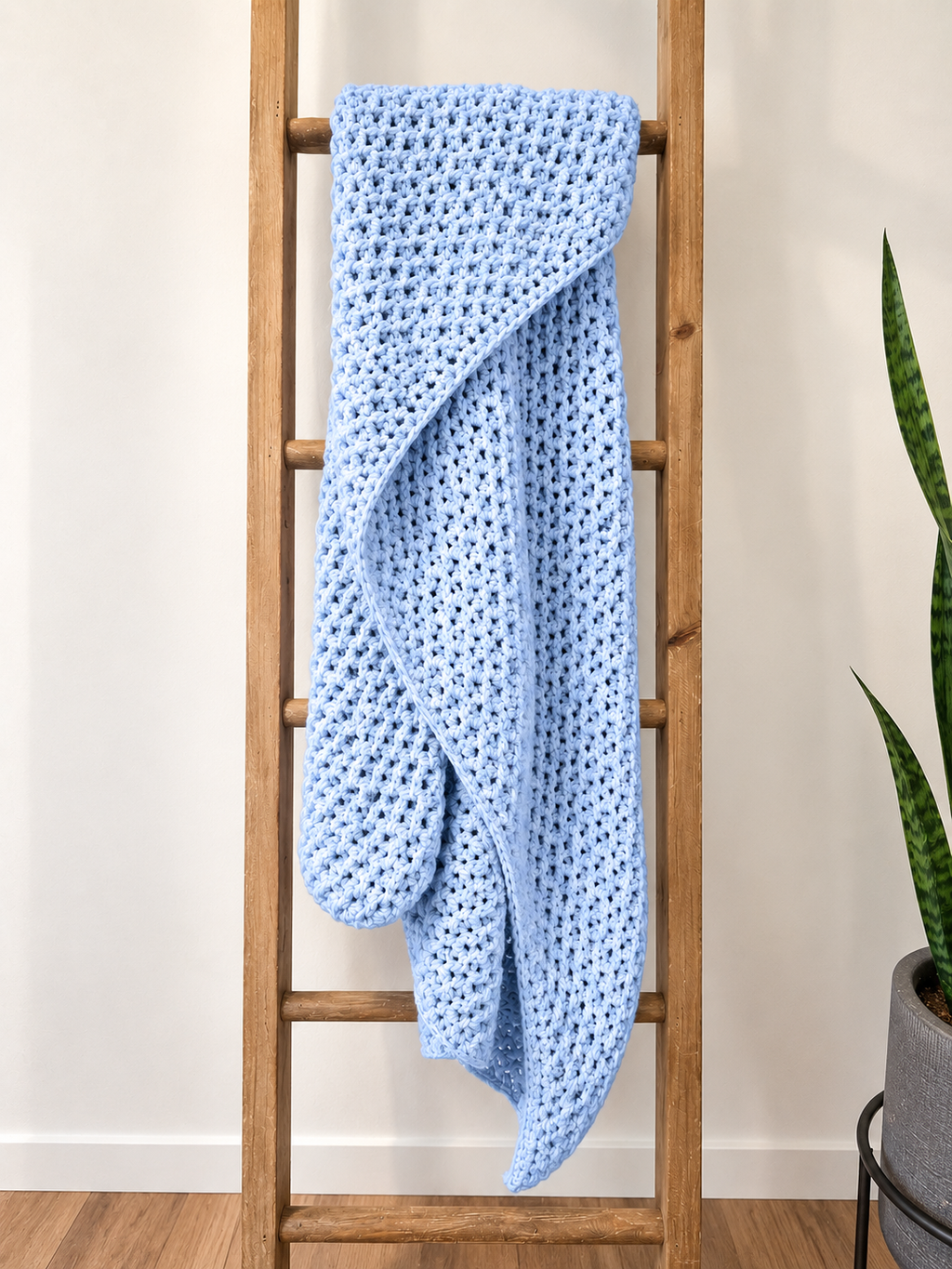 A light blue crocheted blanket with a hood is draped over a wooden ladder in a home setting, with a potted snake plant visible on the right against a white wall.