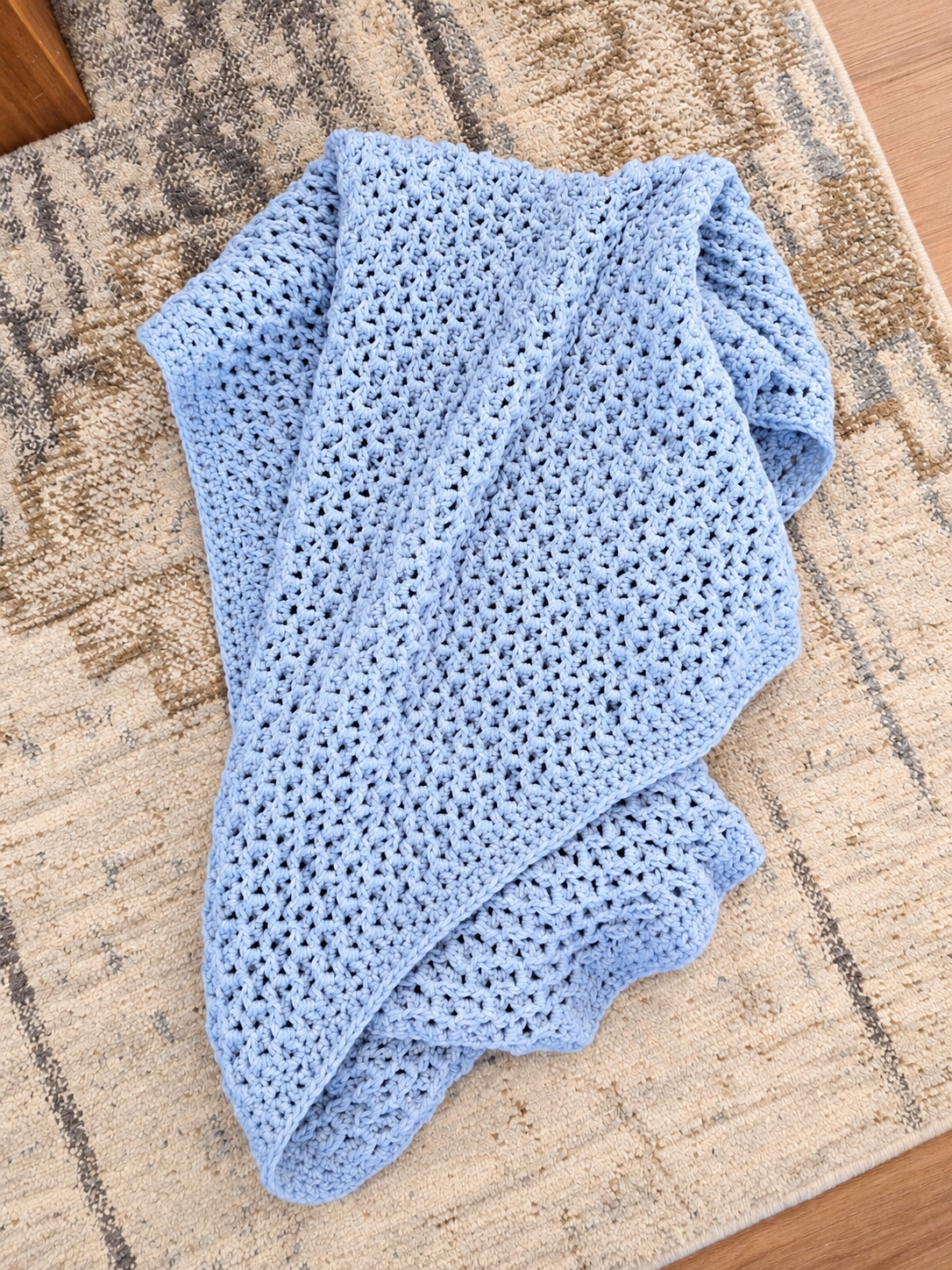 A light blue crocheted throw blanket with an openwork knit pattern, draped on a beige and gray patterned area rug.