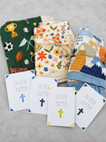 Three folded baby blankets with colorful patterns are arranged on a light gray surface, with four white Baptism confirmation cards in front that read 'ON YOUR BAPTISM' with different colored crosses.