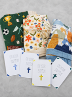 Three folded baby blankets with colorful patterns are arranged on a light gray surface, with four white Baptism confirmation cards in front that read 'ON YOUR BAPTISM' with different colored crosses.