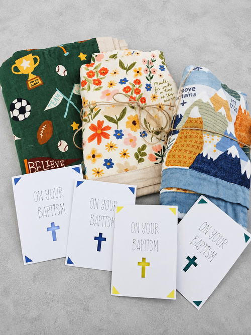 Three folded baby blankets with colorful patterns are arranged on a light gray surface, with four white Baptism confirmation cards in front that read 'ON YOUR BAPTISM' with different colored crosses.