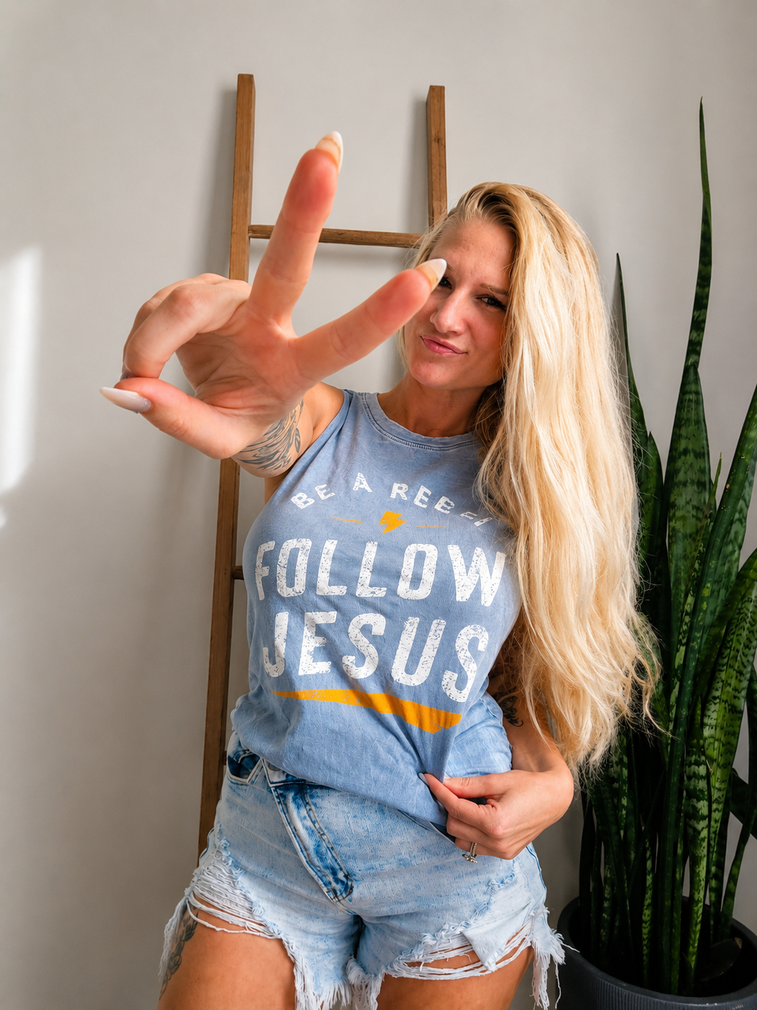 A blonde woman wearing a light blue graphic tank top that reads 'FOLLOW JESUS' in white letters with a yellow stripe, paired with distressed denim shorts. She is posing indoors with a wooden ladder and a green snake plant in the background.