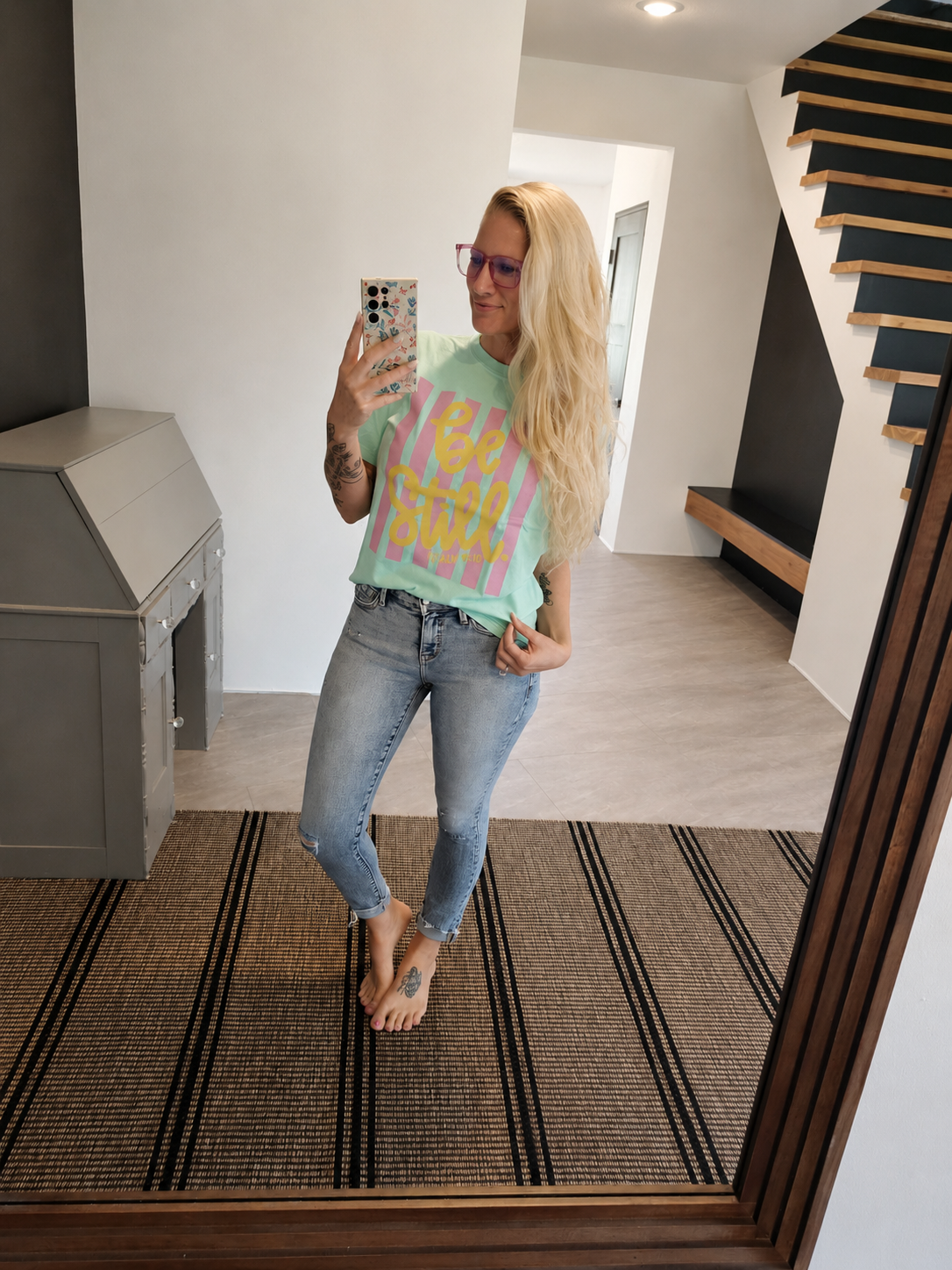 A woman takes a mirror selfie in a modern home interior, wearing a light green graphic t-shirt with pink, yellow, and blue lettering, paired with light-wash blue jeans.