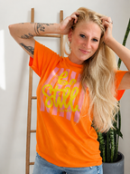 A woman with blonde hair posing in a bright orange graphic t-shirt with pink and yellow text, standing in front of a white wall with a wooden ladder and a potted snake plant.