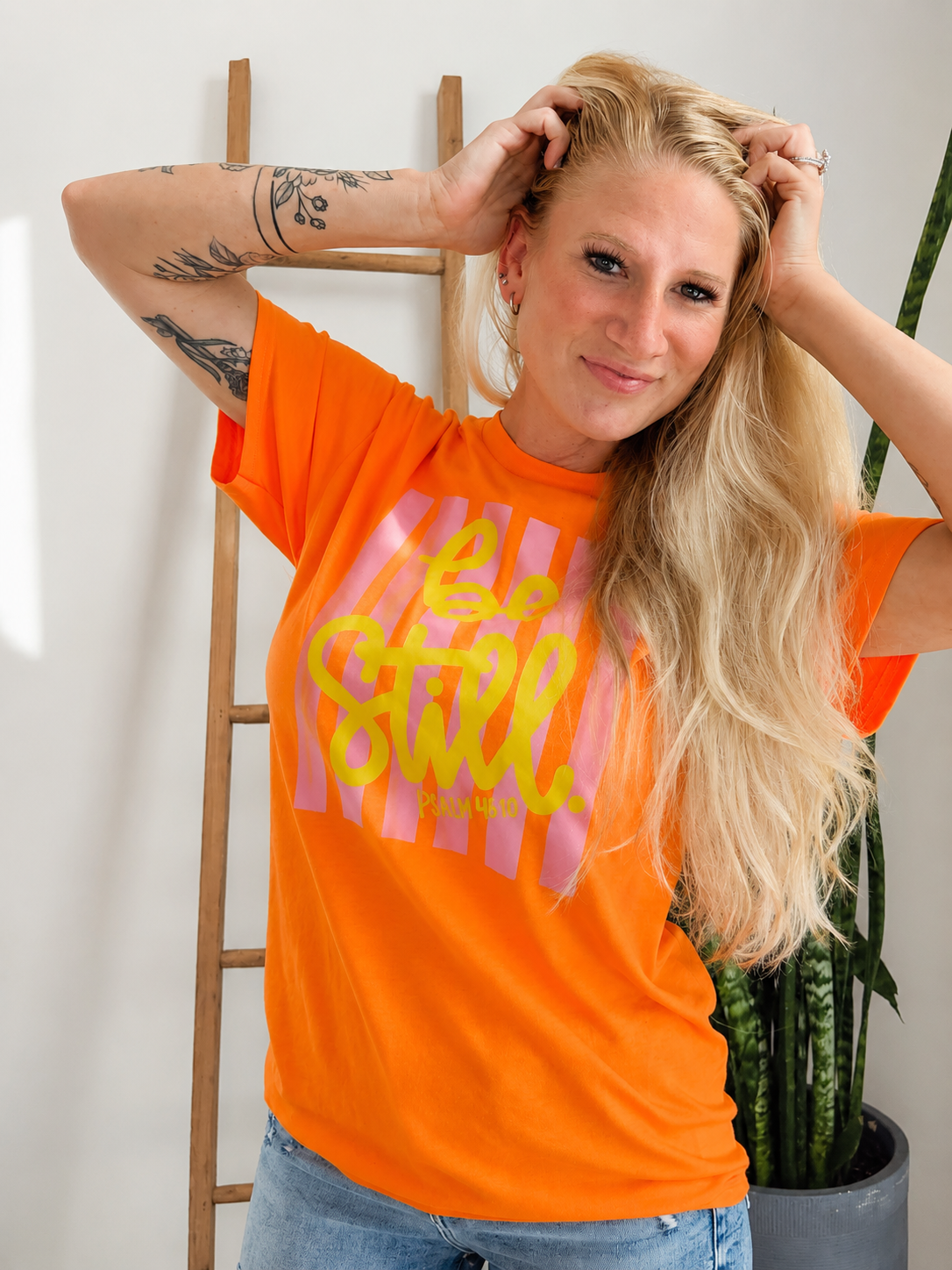 A woman with blonde hair posing in a bright orange graphic t-shirt with pink and yellow text, standing in front of a white wall with a wooden ladder and a potted snake plant.