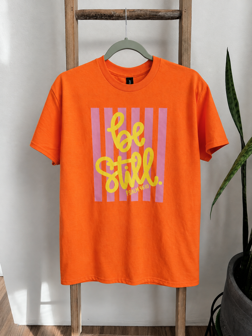 Bright orange short-sleeve t-shirt hanging on a wooden hanger on a rustic wooden ladder, featuring a graphic print with pink vertical stripes and yellow text reading 'be still'.