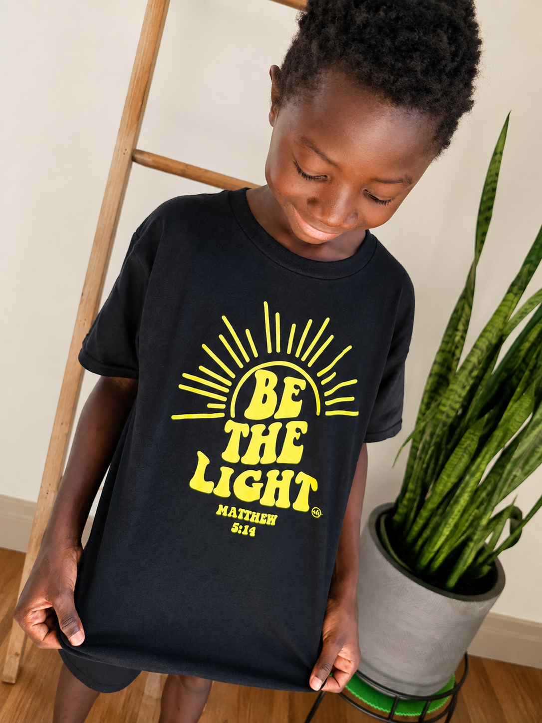 A young boy wearing a black t-shirt with a yellow graphic that reads 'BE THE LIGHT MATTHEW 5:14' under a sunburst design. He is standing in a room with a wooden ladder and a potted snake plant.