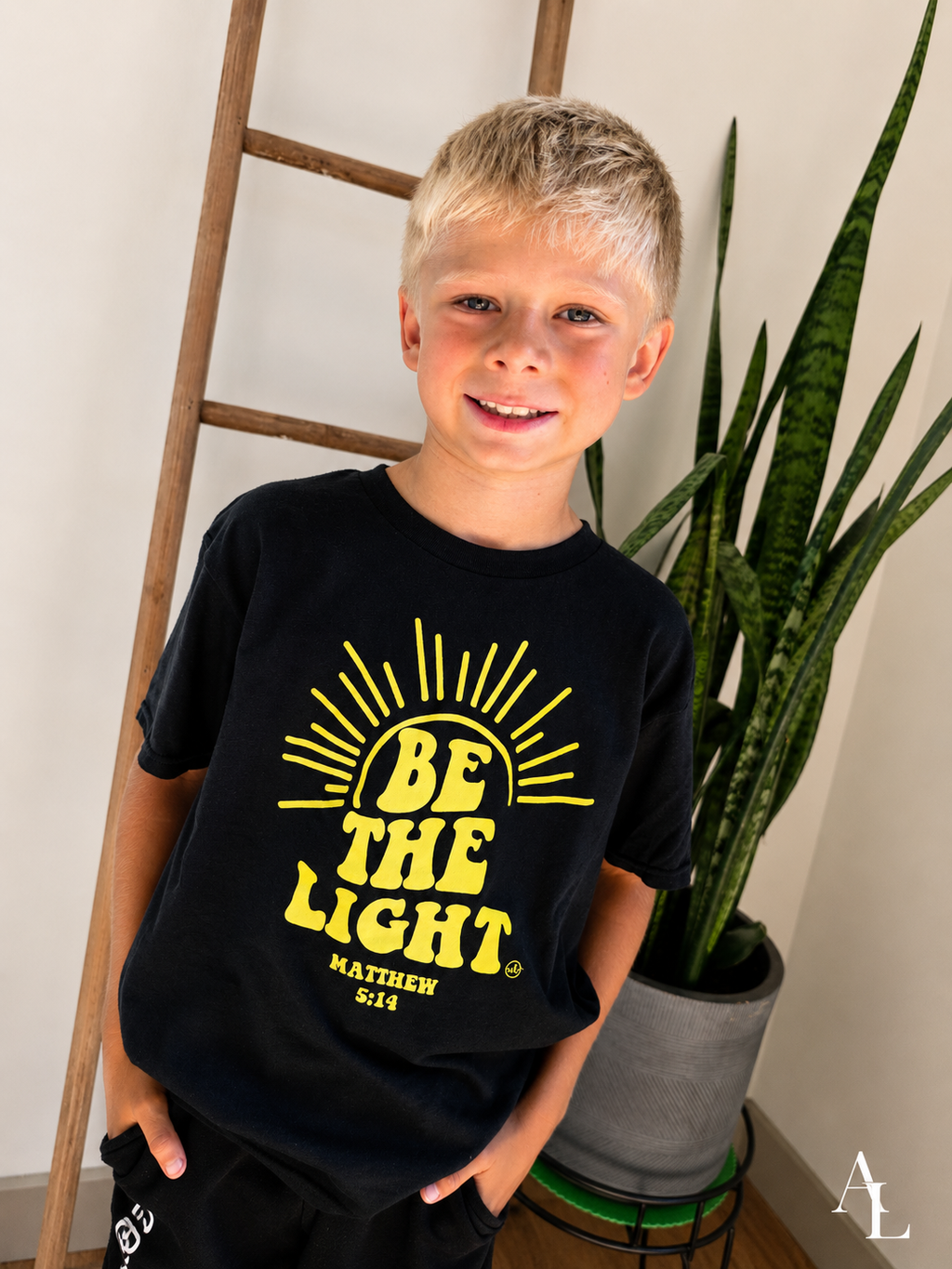 A young boy with blonde hair wearing a black graphic t-shirt that reads 'BE THE LIGHT MATTHEW 5:14' in yellow, standing in a room next to a wooden ladder and a potted snake plant.