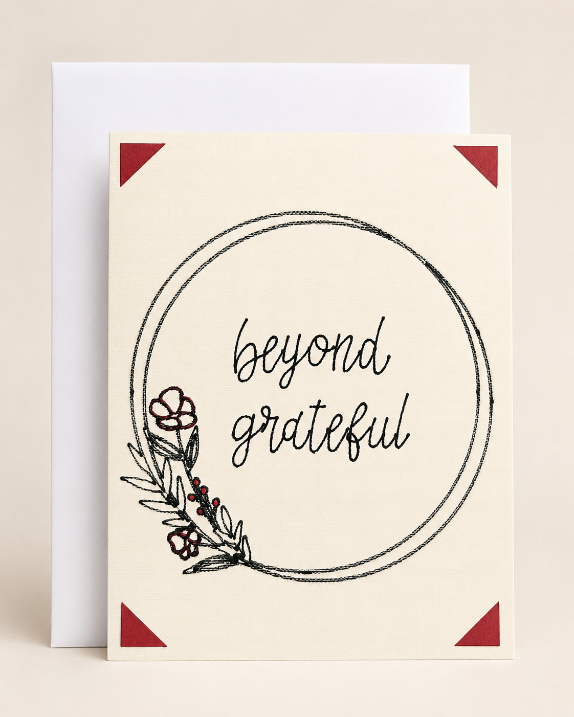 A cream-colored greeting card with the words 'beyond grateful' in black script inside a hand-drawn circular frame, decorated with a small floral illustration in the lower left. The card is shown with a white envelope behind it on a light beige background.