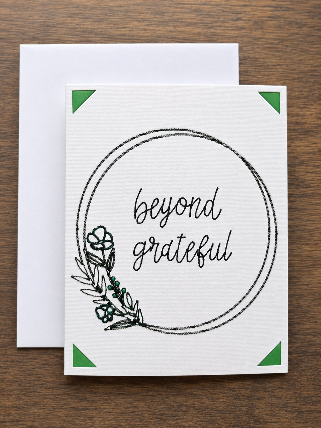 A white greeting card with a black circular border, the words 'beyond grateful' in script, and a small botanical illustration, shown with a white envelope on a brown wood background.