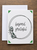 A white greeting card with a black circular border, the words 'beyond grateful' in script, and a small botanical illustration, shown with a white envelope on a brown wood background.