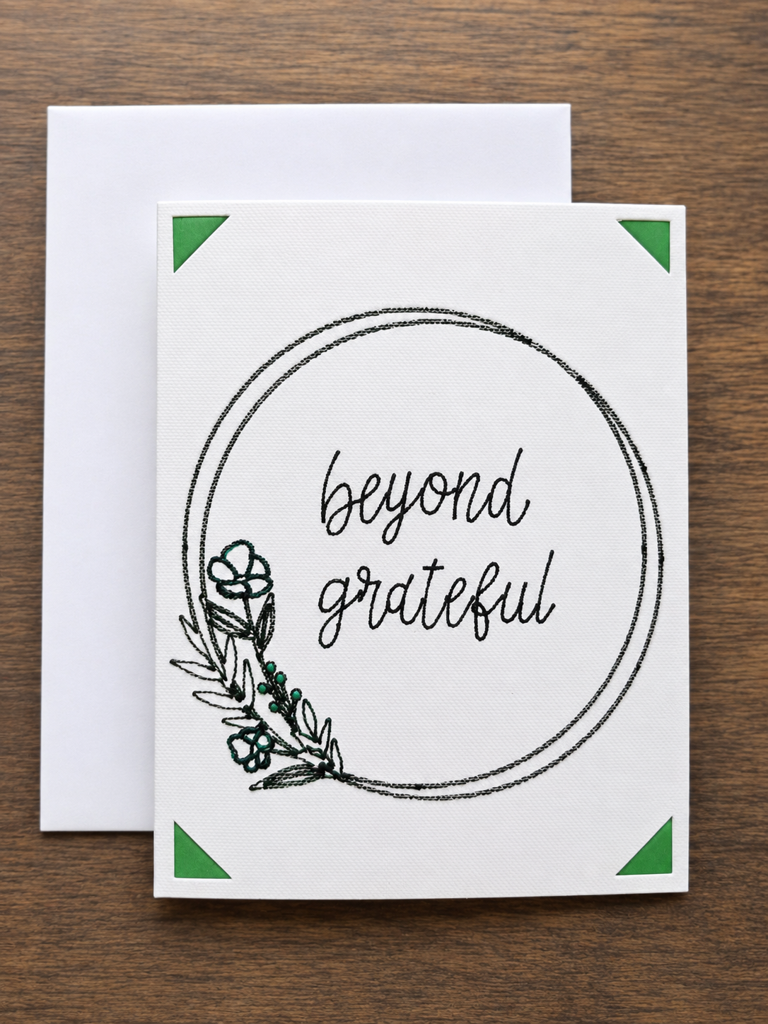 A white greeting card with a black circular border, the words 'beyond grateful' in script, and a small botanical illustration, shown with a white envelope on a brown wood background.