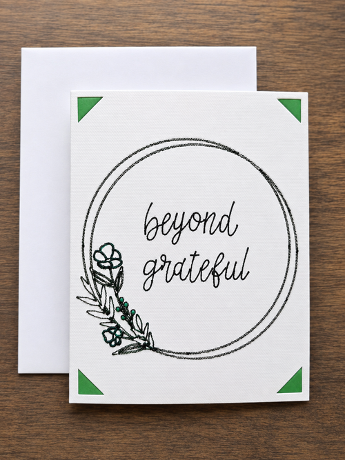 A white greeting card with a black circular border, the words 'beyond grateful' in script, and a small botanical illustration, shown with a white envelope on a brown wood background.