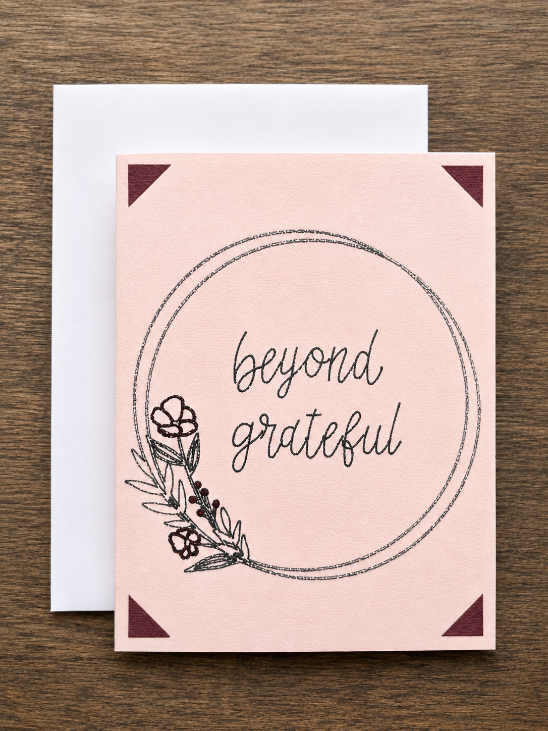 A square greeting card with a pale blush pink background, featuring the words 'beyond grateful' in black script inside a hand-drawn oval, with floral line art in the lower left and dark burgundy corner triangles. A white envelope is partially visible behind it on a brown wood surface.