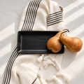A matte black rectangular serving tray styled on a cream-colored woven throw blanket with black stripes and tassels, accompanied by two pears on a light marble surface.
