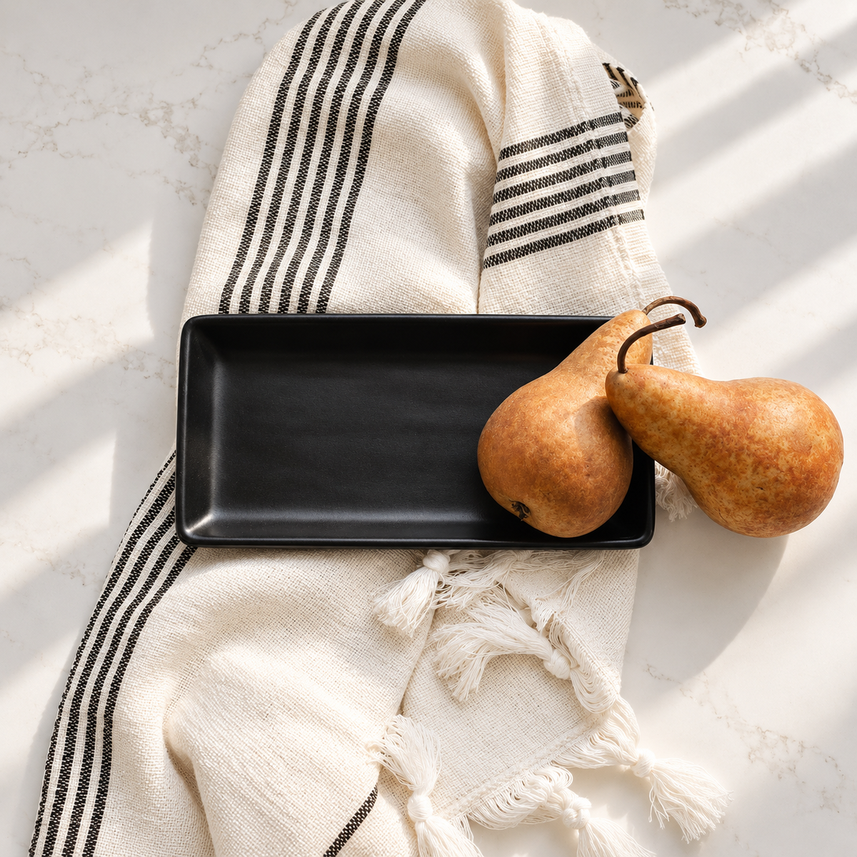 A matte black rectangular serving tray styled on a cream-colored woven throw blanket with black stripes and tassels, accompanied by two pears on a light marble surface.