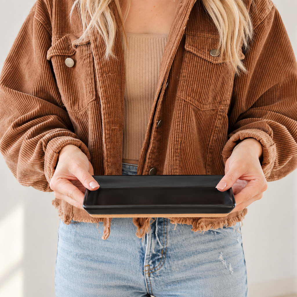 A person wearing a brown corduroy jacket over a beige ribbed top and blue jeans holds a black rectangular tray with a light wood base in front of their torso.