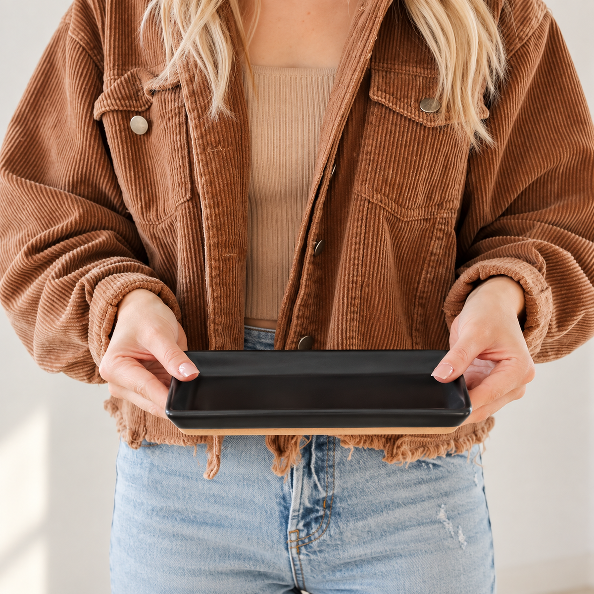 A person wearing a brown corduroy jacket over a beige ribbed top and blue jeans holds a black rectangular tray with a light wood base in front of their torso.