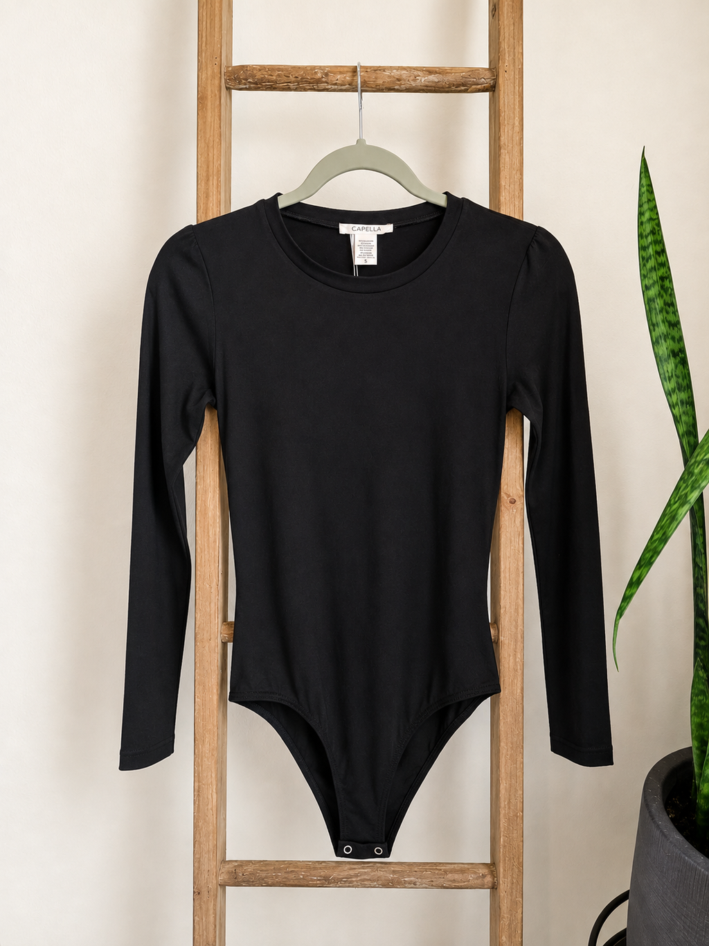 A black long-sleeve bodysuit with a crew neck hangs on a light green hanger from a wooden ladder, with a snake plant visible on the right side.