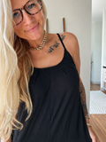 A woman with blonde hair and glasses wearing a black camisole top, gold chain necklace, and gold bracelet, standing in a brightly lit indoor hallway.
