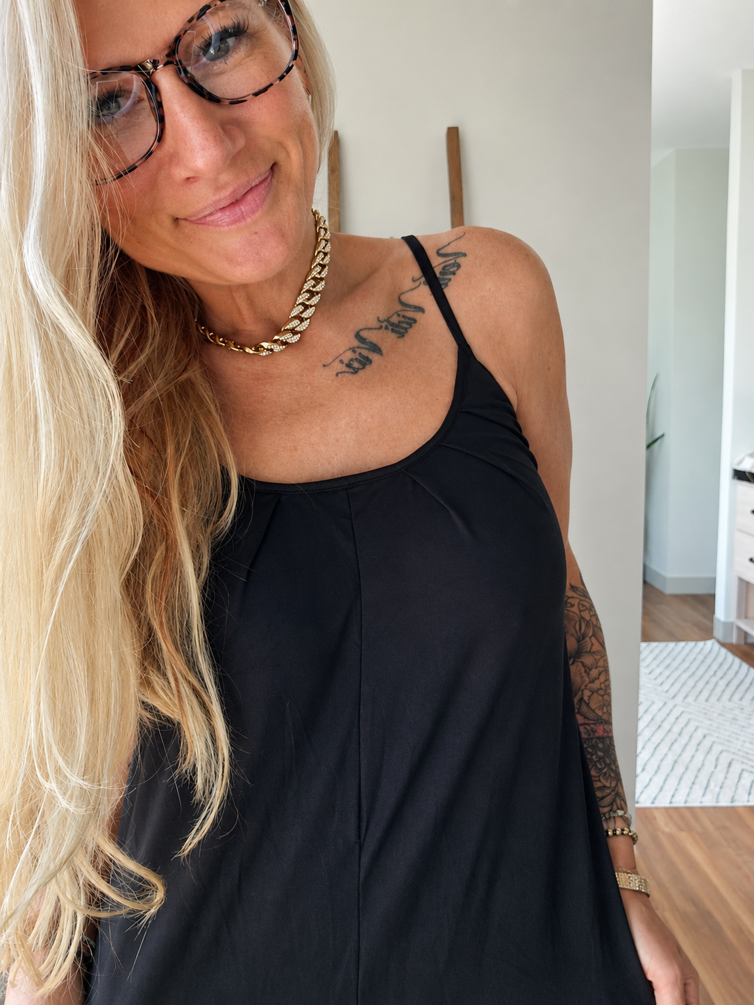 A woman with blonde hair and glasses wearing a black camisole top, gold chain necklace, and gold bracelet, standing in a brightly lit indoor hallway.
