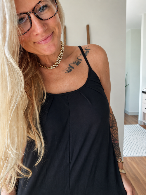 A woman with blonde hair and glasses wearing a black camisole top, gold chain necklace, and gold bracelet, standing in a brightly lit indoor hallway.
