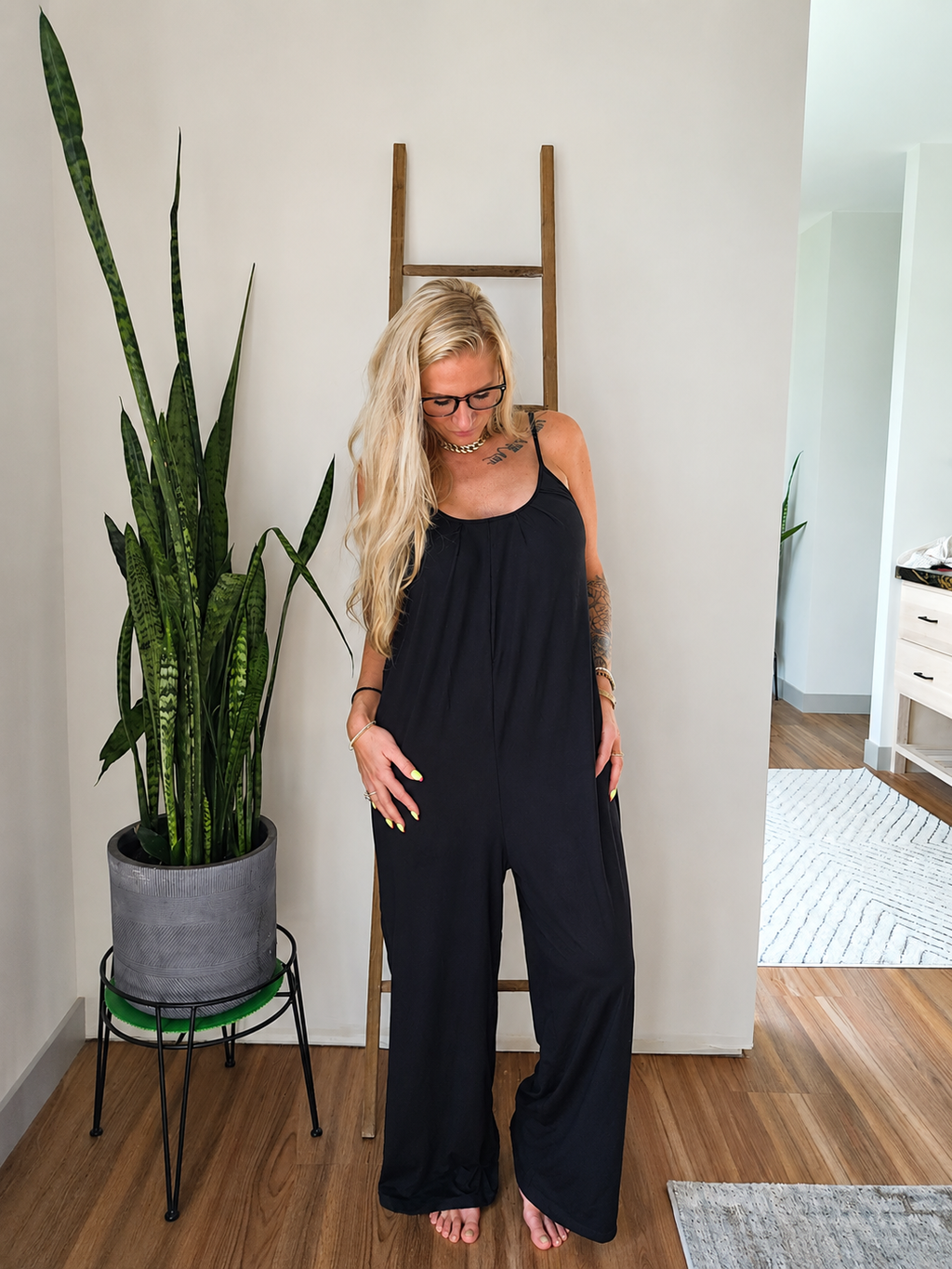 A woman with blonde hair and glasses wearing a black sleeveless jumpsuit, standing barefoot in a room with a wooden floor, a potted snake plant, and a wooden ladder against a white wall.