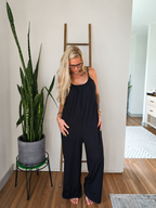 A woman with blonde hair and glasses wearing a black sleeveless jumpsuit, standing barefoot in a room with a wooden floor, a potted snake plant, and a wooden ladder against a white wall.