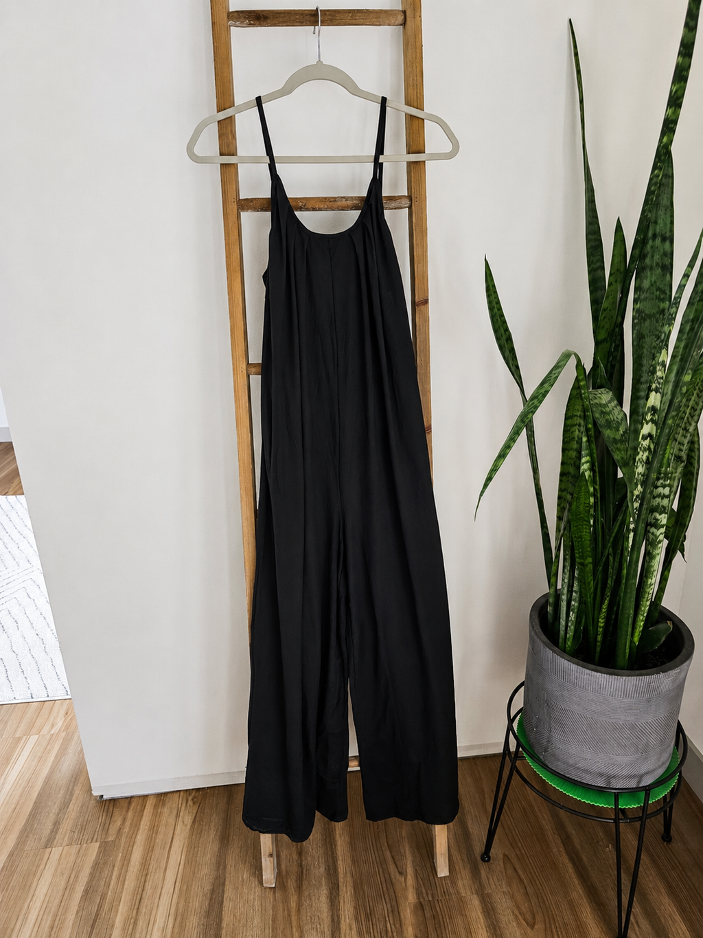A black sleeveless wide-leg jumpsuit hangs on a wooden hanger from a wooden ladder, with a potted snake plant on a stand to the right in a home interior setting.