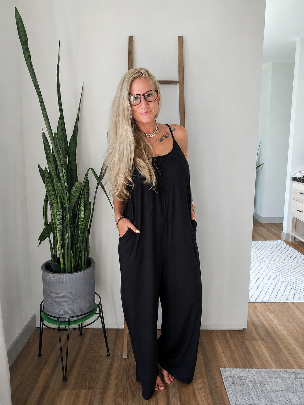 A woman with blonde hair and glasses wearing a black sleeveless jumpsuit with side pockets, standing in a modern room next to a potted snake plant.
