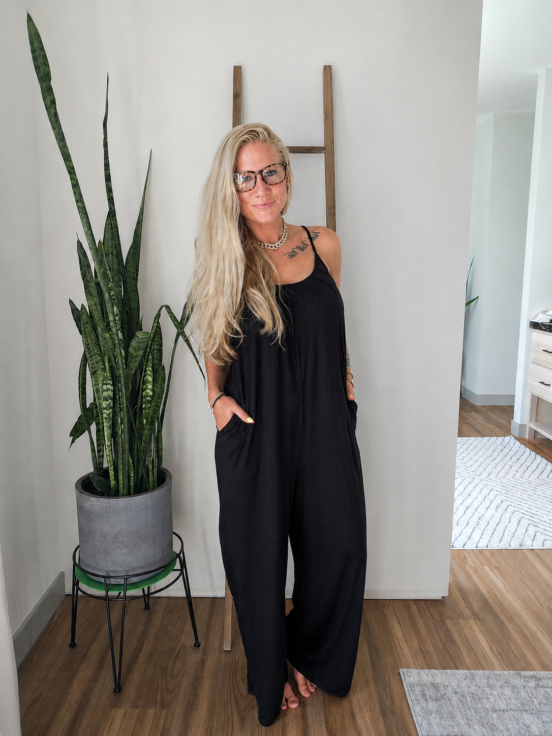 A woman with blonde hair and glasses wearing a black sleeveless jumpsuit with side pockets, standing in a modern room next to a potted snake plant.