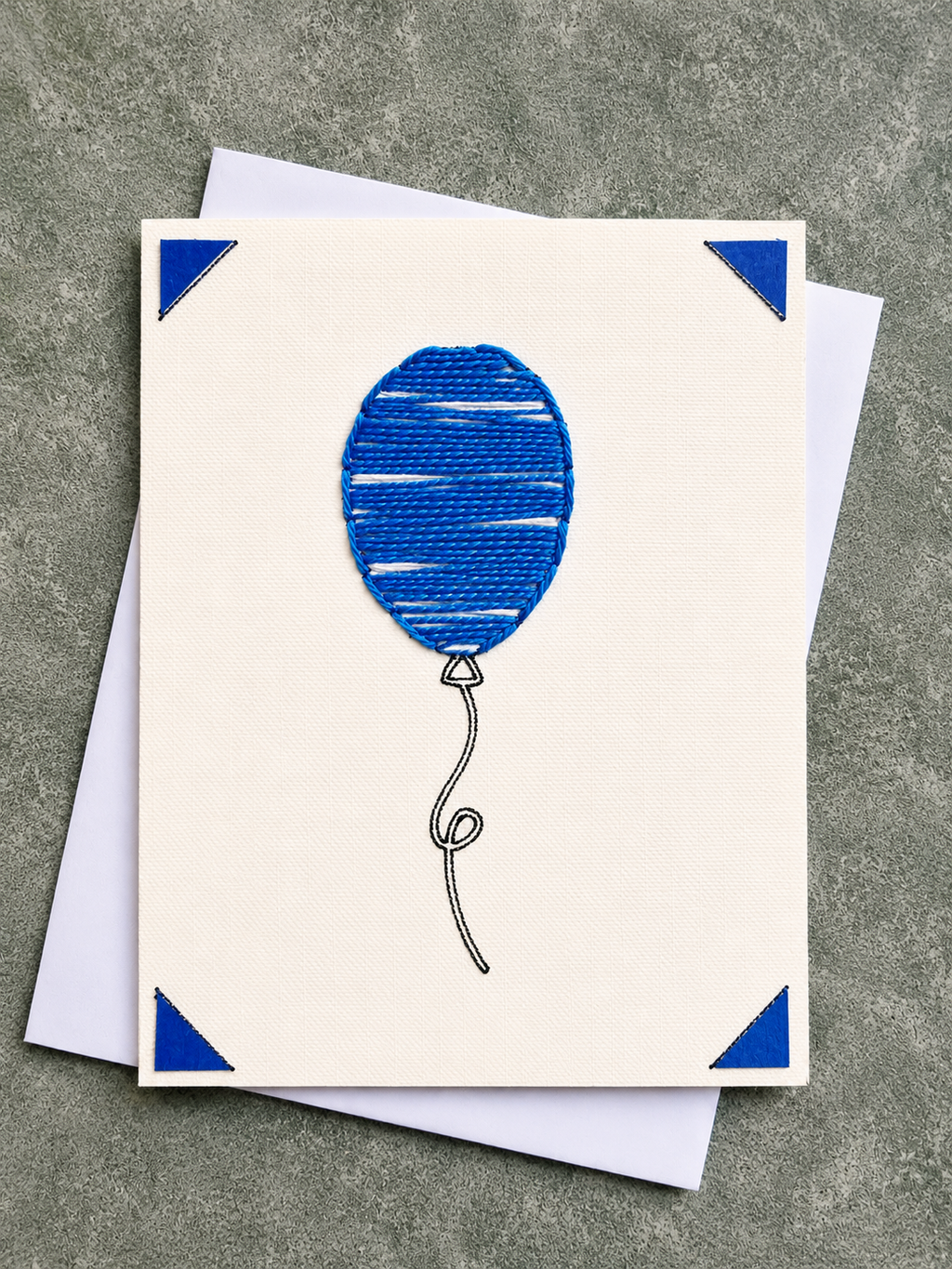 A square cream greeting card featuring a blue balloon illustration with blue corner accents, shown with white envelopes on a textured gray surface.