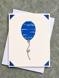 A square cream greeting card featuring a blue balloon illustration with blue corner accents, shown with white envelopes on a textured gray surface.