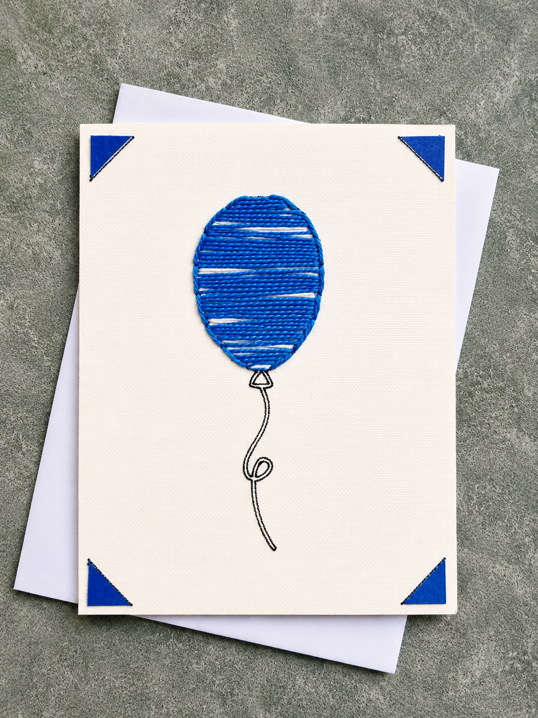A square cream greeting card featuring a blue balloon illustration with blue corner accents, shown with white envelopes on a textured gray surface.