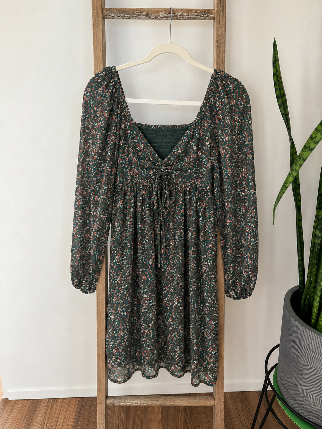 A long-sleeve dark green dress with a dense small floral print in muted red, pink, and beige tones, hanging on a wooden hanger on a rustic wooden ladder. A potted snake plant is visible on the right against a white wall.