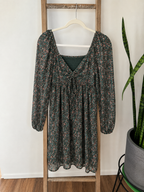 A long-sleeve dark green dress with a dense small floral print in muted red, pink, and beige tones, hanging on a wooden hanger on a rustic wooden ladder. A potted snake plant is visible on the right against a white wall.