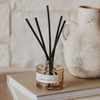 A reed diffuser bottle with black reeds on a beige book, with a white brick wall and a ceramic pitcher in the background. The label reads 'INDICIA WATER REED' and 'Cinnamon and Vanilla'.