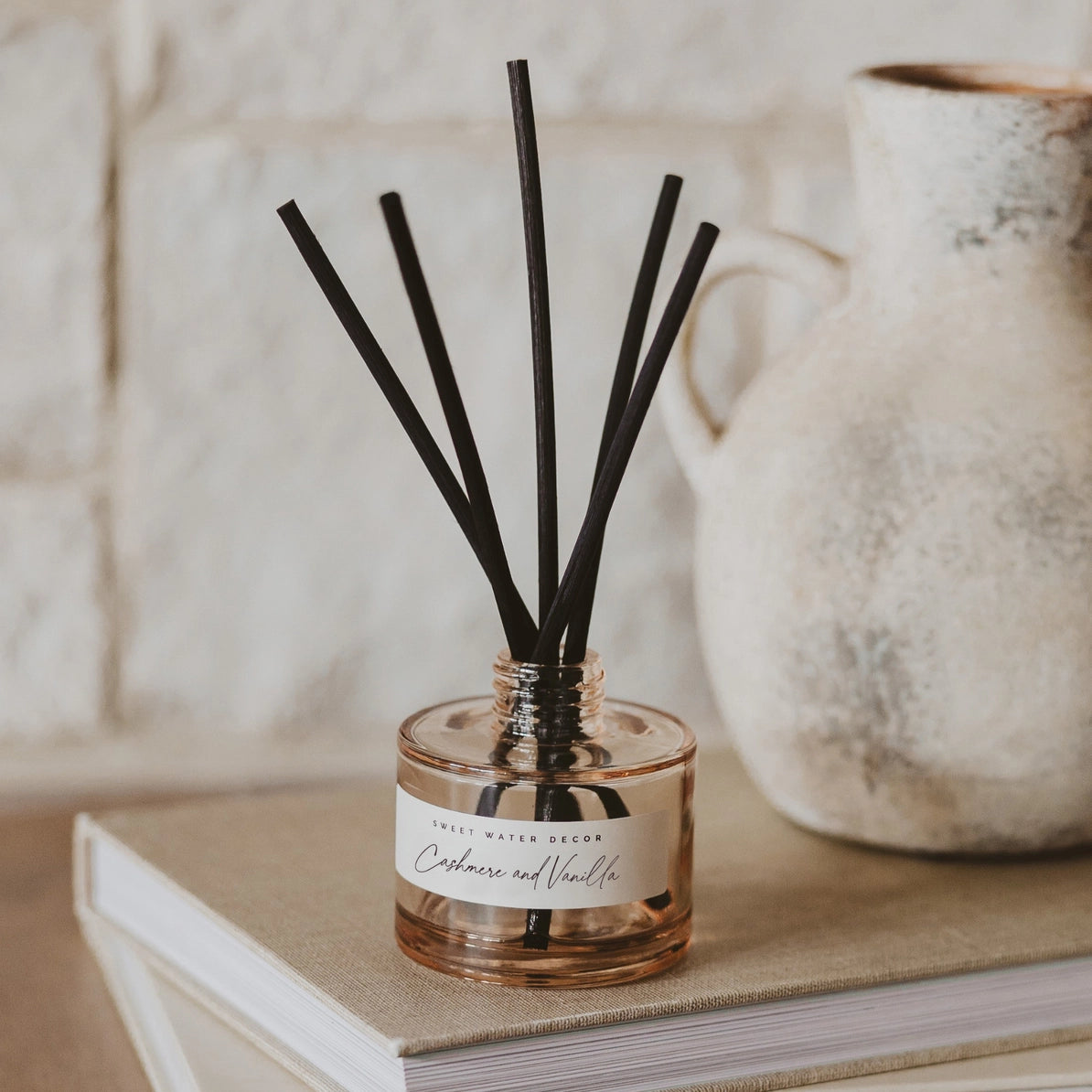 A reed diffuser bottle with black reeds on a beige book, with a white brick wall and a ceramic pitcher in the background. The label reads 'INDICIA WATER REED' and 'Cinnamon and Vanilla'.